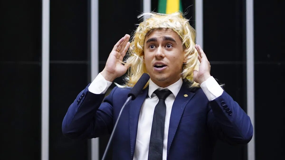 Nikolas Ferreira, deputado federal, usando peruca loira, com as mãos levantadas próximas à cabeça, discursando à tribuna da Câmara dos Deputados