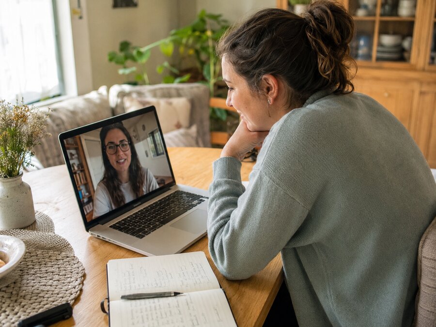 Mulher em uma videochamada genuína em casa, ouvindo com atenção, caderno de anotações aberto ao lado