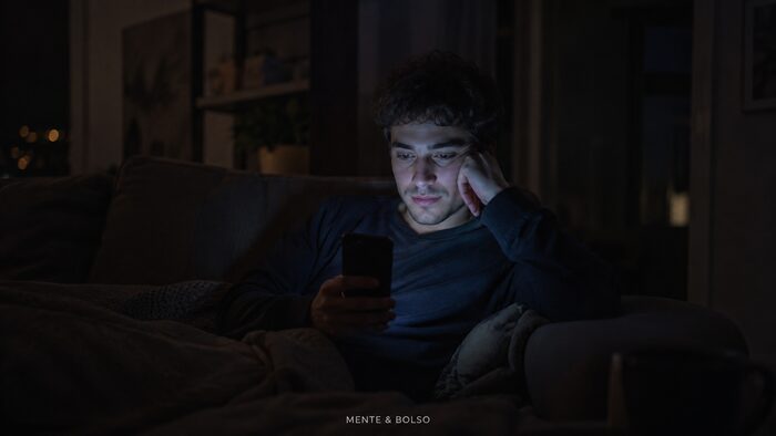 Homem sozinho no sofá à noite, iluminado apenas pela luz azulada do celular em um cômodo escuro, com expressão pensativa e cansada