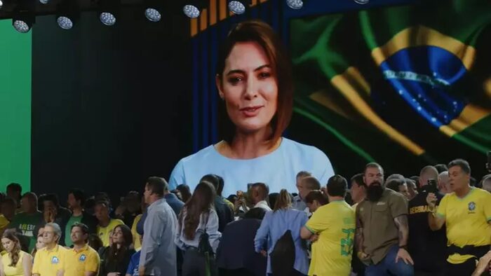 Telão exibindo o rosto de Michelle Bolsonaro durante a convenção do PL em Brasília, com bandeira do Brasil ao fundo e militantes na plateia