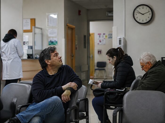 Sala de espera de hospital com pessoas aguardando atendimento, um homem olhando preocupado para o relógio na parede