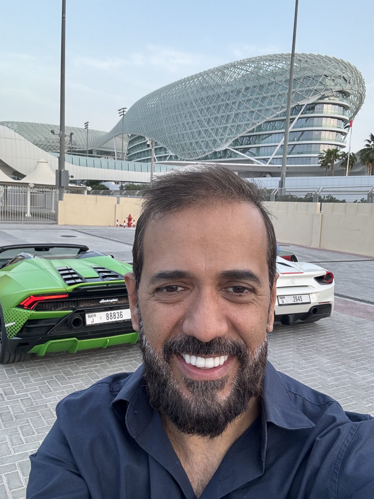 Joaquim sorrindo em frente ao autódromo Yas Marina, em Abu Dhabi, com uma Lamborghini verde e uma Ferrari branca ao fundo